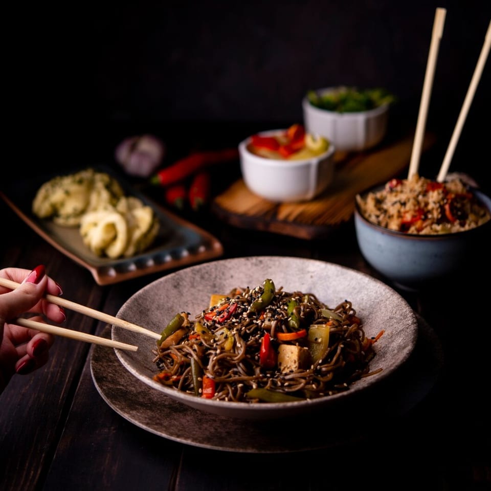 A hand with red nails uses chopsticks to pick up soba noodles with vegetables and tofu.
