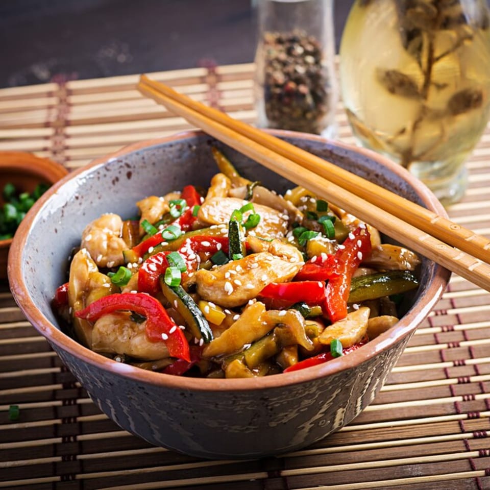A bowl of TERIYAKING chicken stir-fry with red peppers, zucchini, and sesame seeds, with chopsticks.