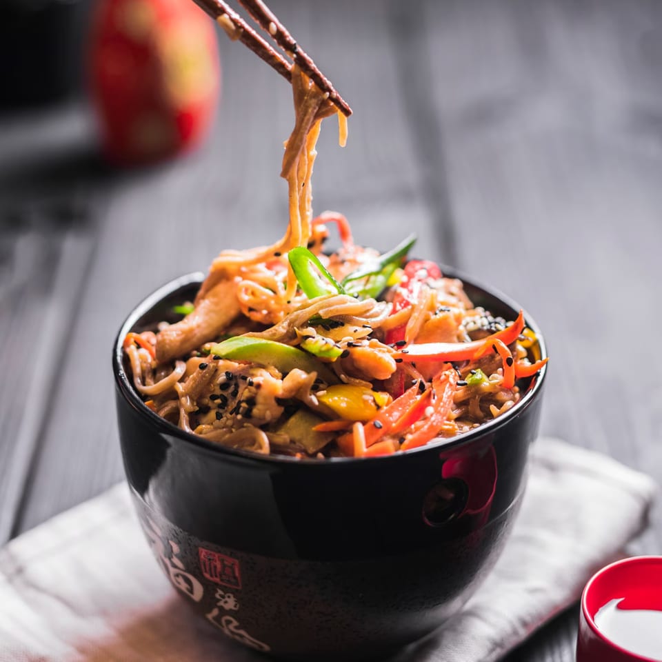 Chopsticks lifting noodles from a black bowl of TERIYAKING stir-fry with vegetables and sesame seeds.